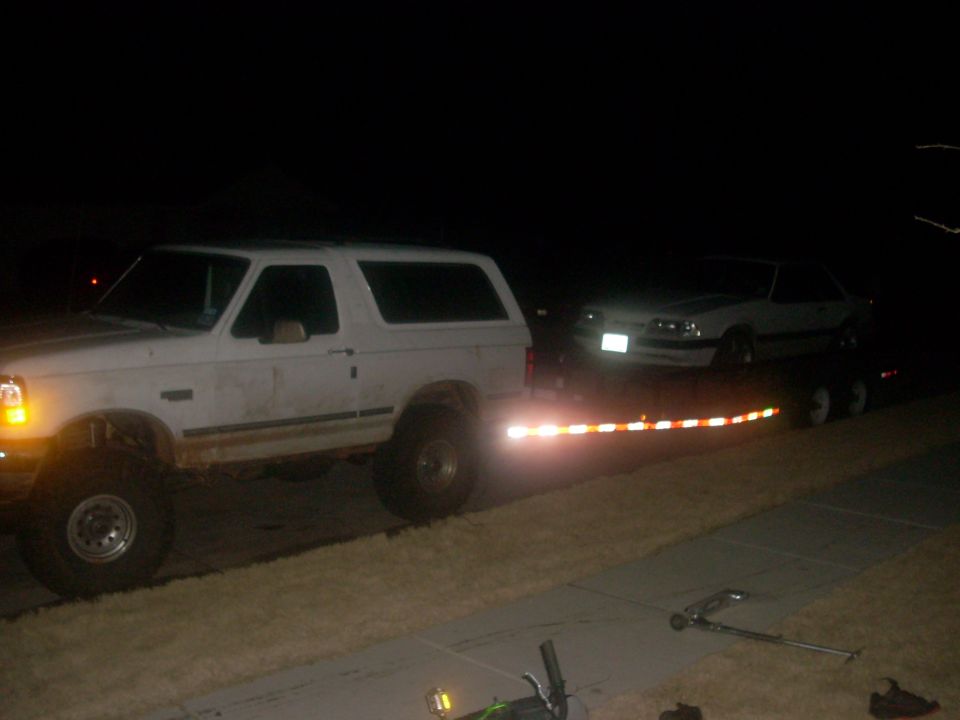 Lifted white Ford Bronco towing a trailer with a white Fox-body Mustang at night