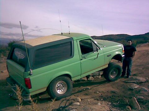 1996 Ford Bronco - A green 1996 Ford Bronco with a tan roof is parked off-road, with a man standing beside it.