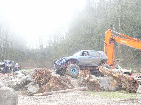 Modified off-road SUV climbing over logs and rocks, with an excavator in the background