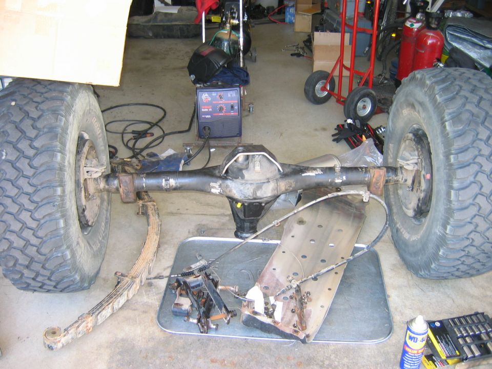 rear axle assembly with large tires, leaf springs, and brake components, next to a welding machine