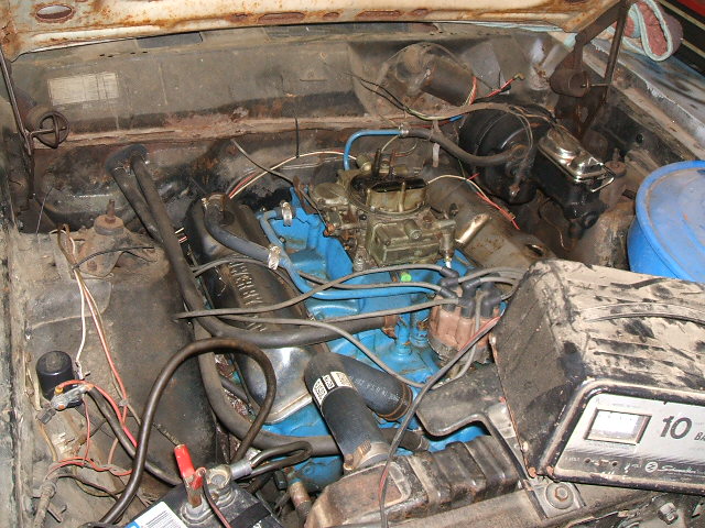 Engine bay with blue engine block, carburetor, and battery. Some rust visible
