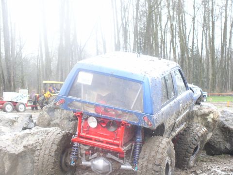 Rear view of blue off-road SUV with exposed red frame and large tires stuck in mud