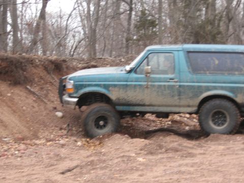 1996 Ford Bronco - A teal SUV navigates a muddy, off-road terrain with trees in the background.