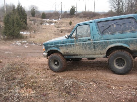 1996 Ford Bronco - A teal 1996 Ford Bronco is seen from the side, heavily coated in mud, driving on a dirt path with sparse vegetation and power lines in the…