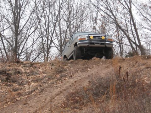 1996 Ford Bronco - A light blue 1996 Ford Bronco navigates an off-road incline amidst bare trees.