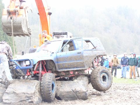 Muddy SUV with lifted suspension and large tires is being lifted by an excavator. People watch in the backgrou