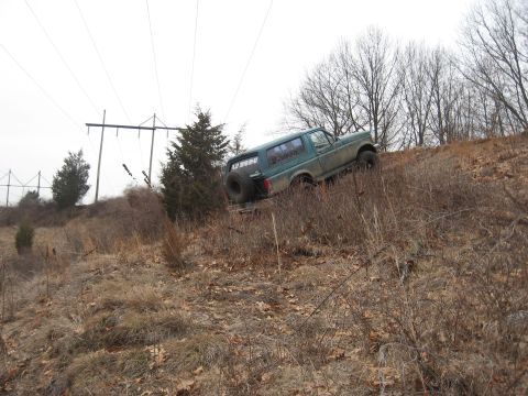 1996 Ford Bronco - A green 1996 Ford Bronco navigates a steep, grassy incline under a cloudy sky, with power lines visible in the background.