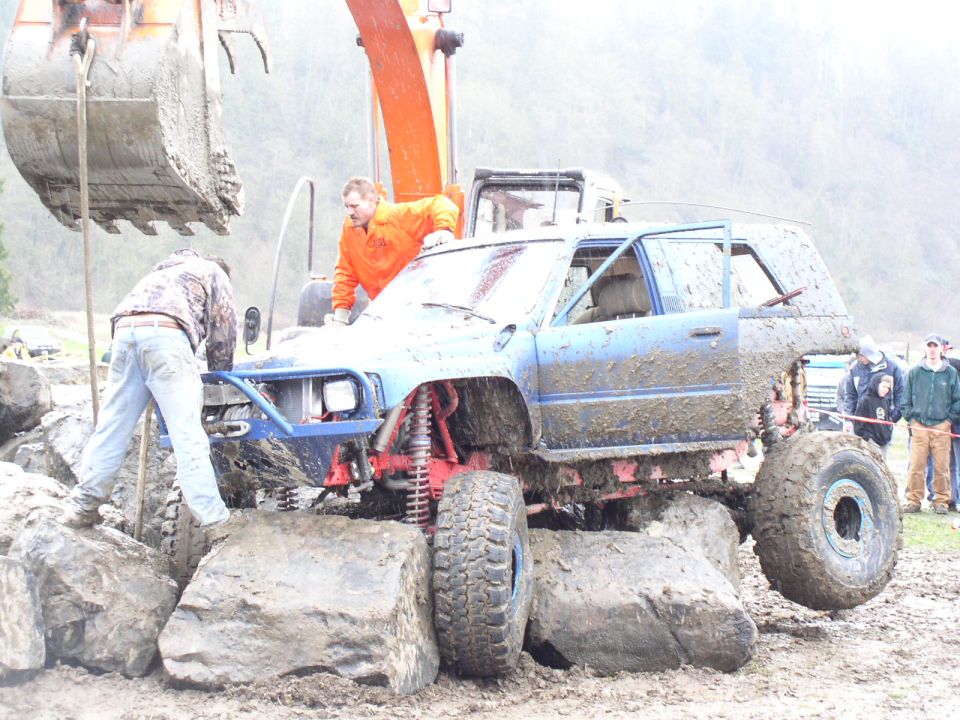 Mud-covered blue SUV with oversized tires on rocks, front bumper removed, driver's door open