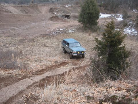 1996 Ford Bronco - A teal SUV navigates a dirt track in a rural, wooded area with some snow patches visible.