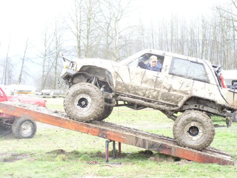 Mud-covered SUV with large tires on a wooden ramp, driver visible