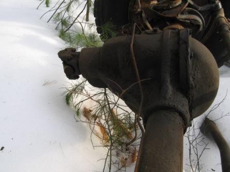 1993 Ford Bronco - Close-up of a rusty differential housing and driveshaft, partially covered in snow and pine needles.