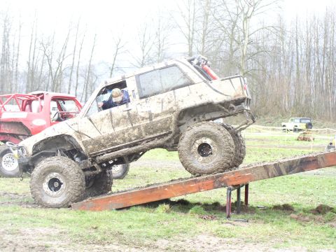 Mud-covered off-road SUV on a wooden ramp, with a red truck behind it