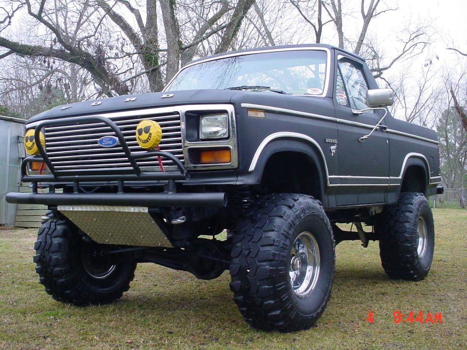 Black lifted Ford truck with brush guard, big mud tires, and yellow smiley light covers on grass