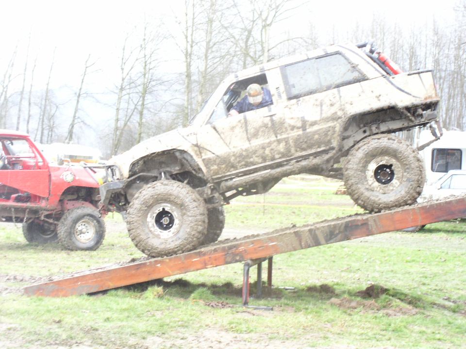 Muddy off-road vehicle climbing a ramp, with a red truck nearby