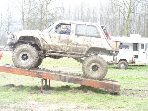 Muddy off-road vehicle with large tires on a wooden ramp; a white trailer is parked behind it
