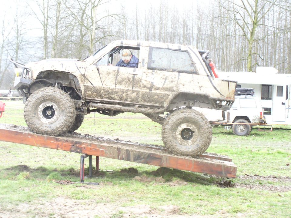 Muddy off-road vehicle with large tires on a wooden ramp; a white trailer is parked behind it
