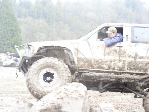 Muddy off-road vehicle with large tires, driver wearing a cap and jacket, navigating rough terrain