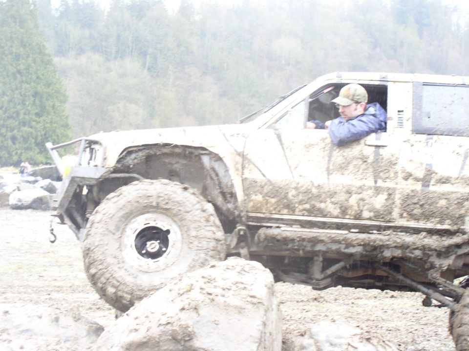 Muddy off-road vehicle with large tires, driver wearing a cap and jacket, navigating rough terrain
