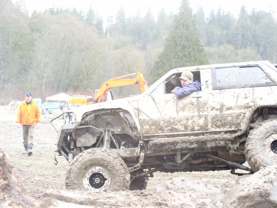 Mud-caked off-road SUV with exposed suspension, driver in window, parked in mud