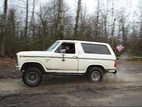 1980 Ford Bronco - A white 1980 Ford Bronco kicks up mud as it drives through a wooded area.