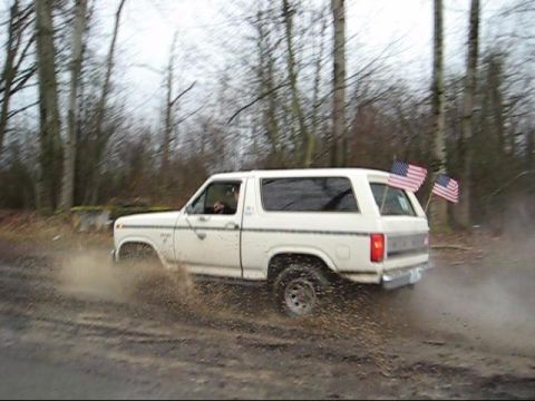 1980 Ford Bronco - A white 1980 Ford Bronco kicks up mud as it drives through a wooded area.