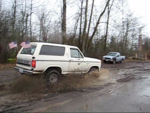 1980 Ford Bronco - A white 1980 Ford Bronco navigates a muddy off-road path, kicking up water and dirt.
