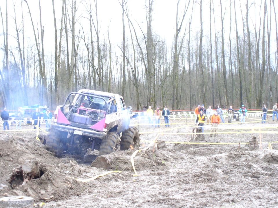 Rear view of a modified off-road truck with a roll cage stuck in mud, emitting smoke