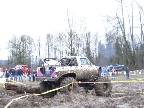 Mud bogging truck stuck in mud with roll cage and large tires. Spectators watch in the background