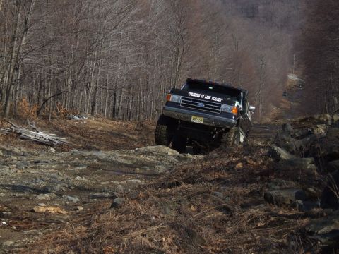 1976 Toyota Land Cruiser - Black Ford F-Series truck navigates a challenging rocky off-road trail in a wooded area.