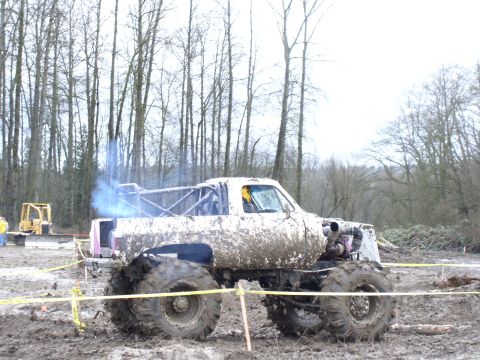 Mud-covered monster truck with large tires and exposed engine, emitting blue smoke from exhaust