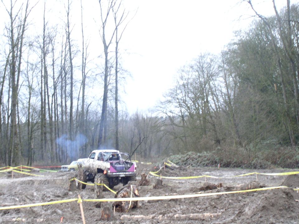 Purple mud truck spinning through a roped-off bog course trailing exhaust smoke, bare trees behind