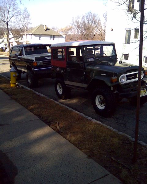 1989 Ford Bronco - Two classic off-road SUVs, a Toyota Land Cruiser and a Ford Bronco, are parked on a residential street.