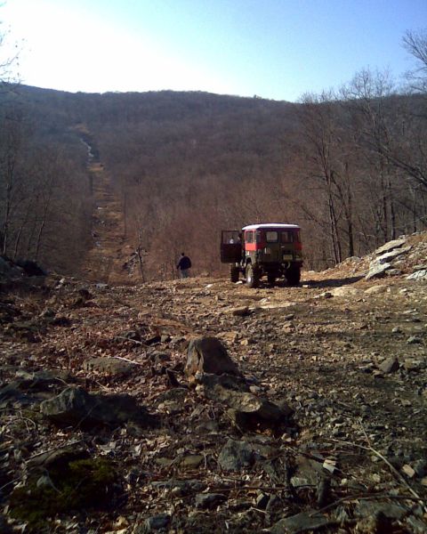 1989 Ford Bronco - A person stands near an off-road vehicle on a steep, rocky incline with trees in the background.