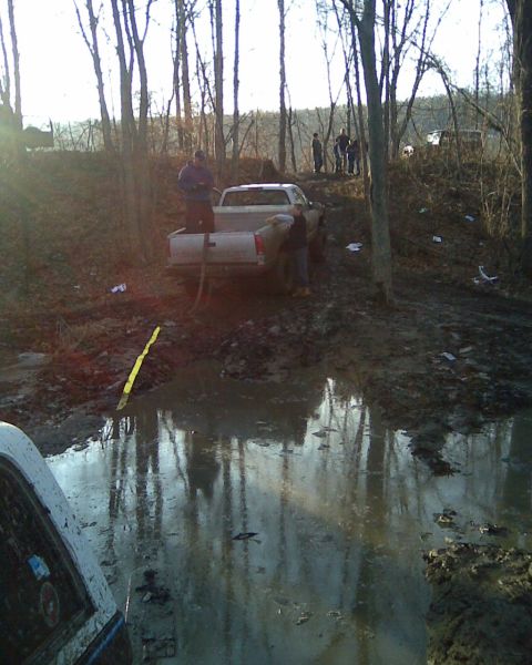 1989 Ford Bronco - A truck is stuck in a muddy, wooded area, with people attempting to pull it out with a tow strap.