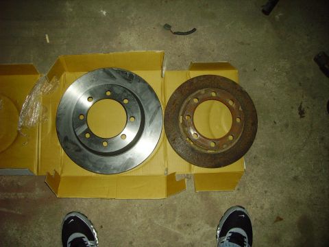 1991 Ford Bronco - A comparison of a new brake rotor with a heavily rusted old one, laid out on a concrete floor.