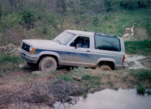 White two-door Ford SUV with blue stripes parked on a muddy off-road trail near water