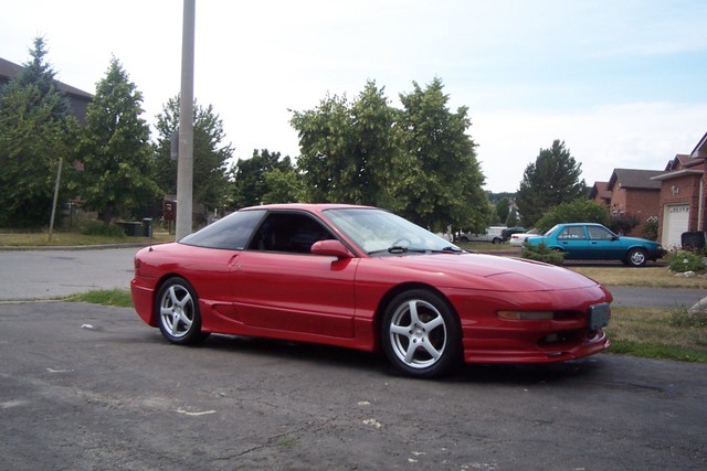 red 1995 Ford Probe parked on asphalt, houses and trees in background, blue sedan parked across street