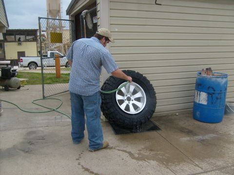 1989 Ford Bronco - A man cleans a large tire and wheel assembly with a garden hose in front of a beige sided building.