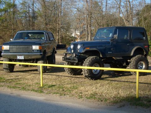 1989 Ford Bronco - Two , lifted vehicles, a Ford Bronco and a Jeep, are parked outdoors on a sunny day.