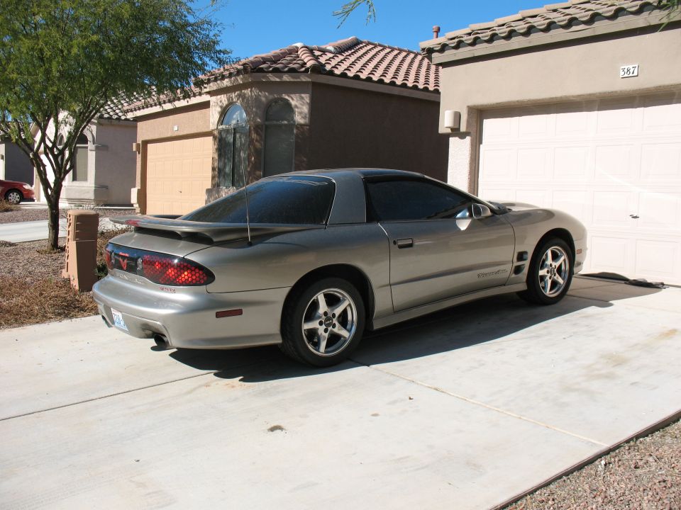 Pewter Pontiac Trans Am coupe on a driveway, driver-side rear 3/4 view with rear wing