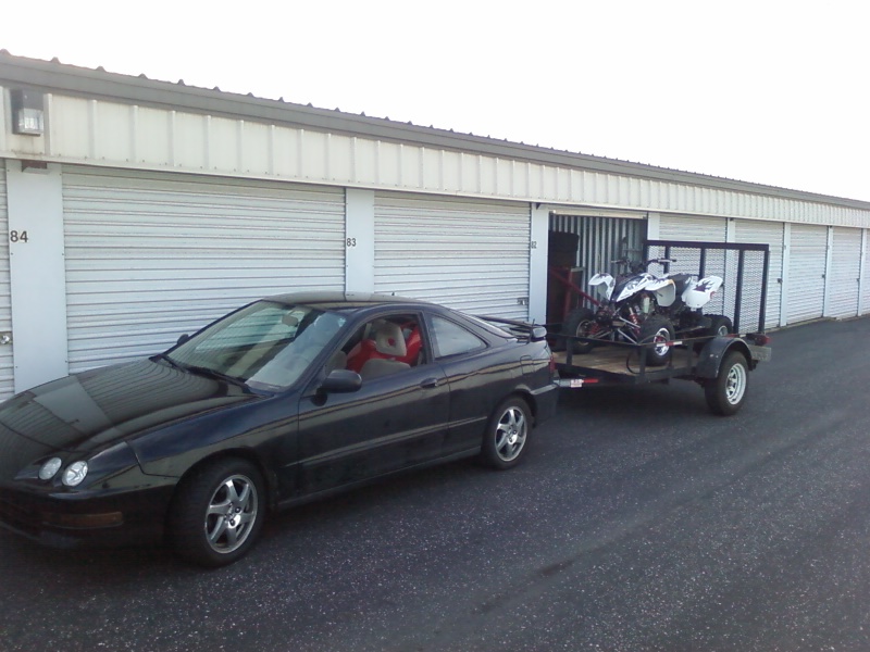 Black coupe towing a trailer with two ATVs, parked in front of storage units