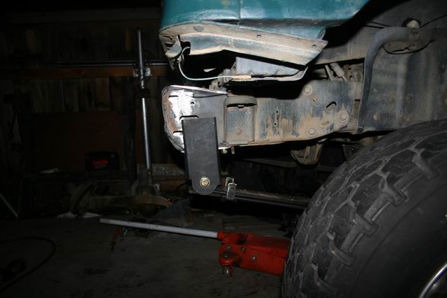 Close-up of a custom bracket being bolted to a vehicle's frame, likely for a bumper modification. A red hydraulic jack is visible underneath