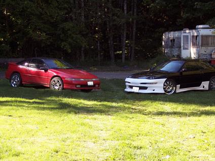 1991 Mitsubishi Eclipse - Two sporty coupes are parked on a grassy area with trees and a trailer in the background.