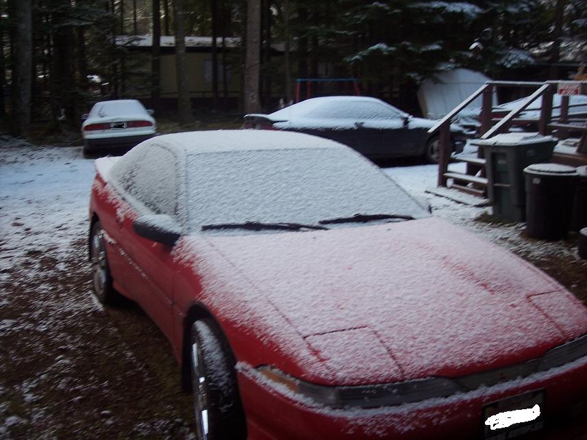 Red coupe with snow on its roof and hood, parked outside. The front end is visible, showing pop-up headlights
