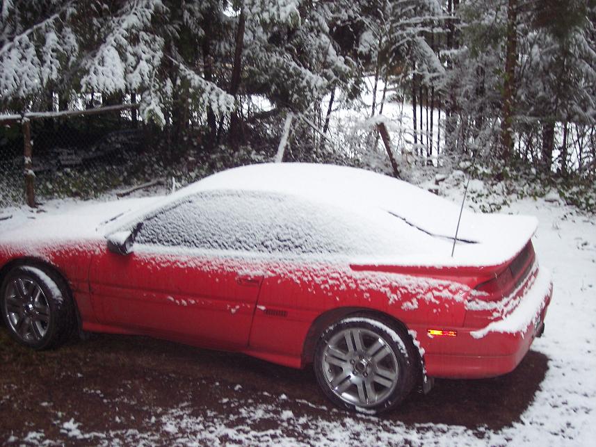 Red sports car covered in snow, parked outside with trees in the background