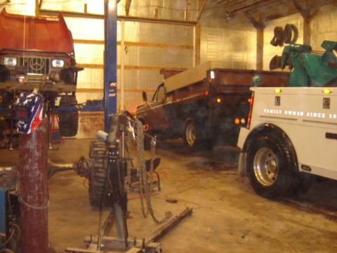 1992 Ford Bronco - A dump truck and a tow truck are parked inside a garage, with another vehicle on a lift in the background.