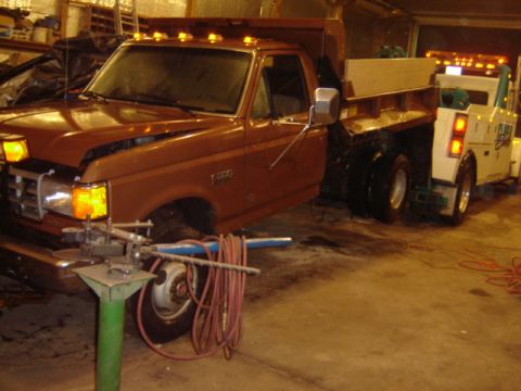 1992 Ford Bronco - A brown Ford F-Series dump truck, likely from the late 1980s, is being towed by a white tow truck inside a garage.