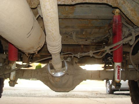 1990 Ford Bronco - Undercarriage view showing the differential, driveshaft, and red Rancho shock absorbers. A second vehicle is visible in the background.