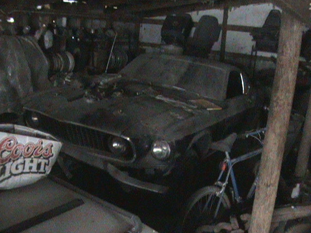 old car with a grille that looks like a 1969 Mustang, parked inside a cluttered garage with a bike and some seats