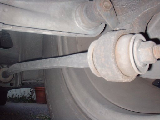 Underbody close-up of a suspension link with rusty bushing and nut beside a tire